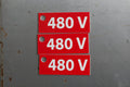 A red rectangular electrical sign with white text reading '480 V' and a mounting hole on the left, shown against a textured brown stone background.