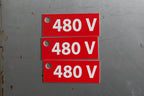 A red rectangular electrical sign with white text reading '480 V' and a mounting hole on the left, shown against a textured brown stone background.