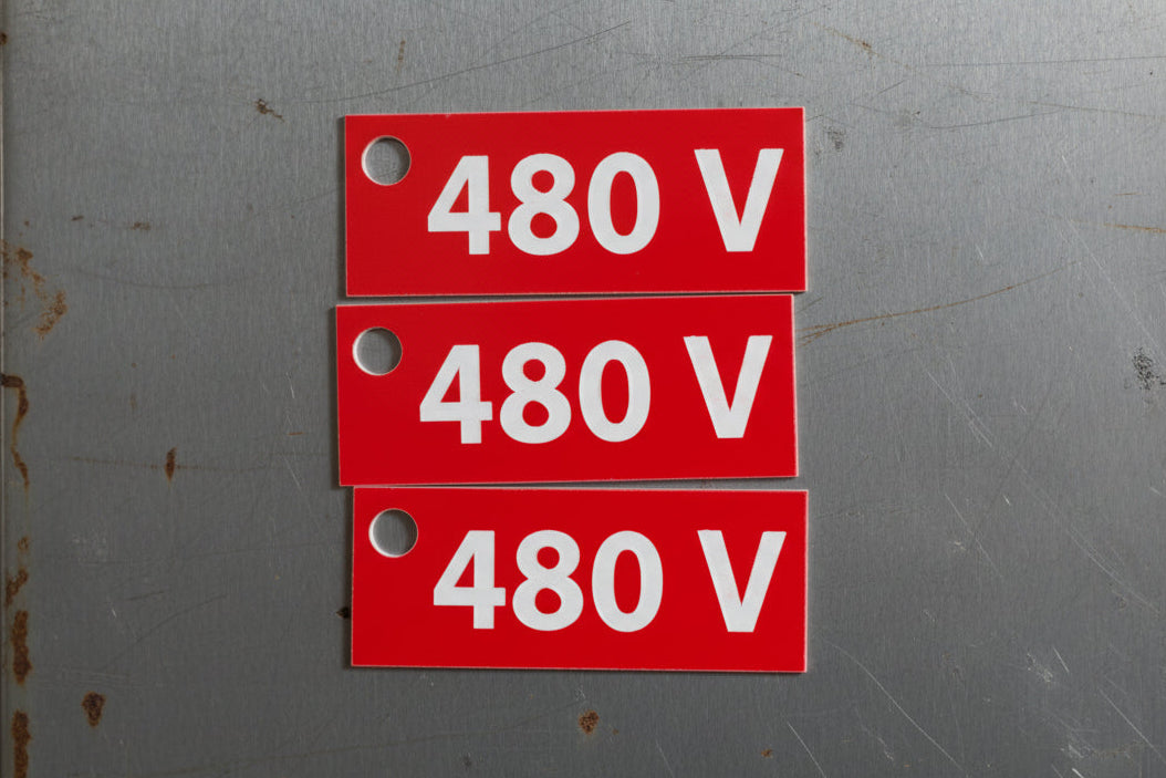 A red rectangular electrical sign with white text reading '480 V' and a mounting hole on the left, shown against a textured brown stone background.