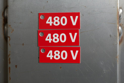 A red rectangular electrical sign with white text reading '480 V' and a mounting hole on the left, shown against a textured brown stone background.